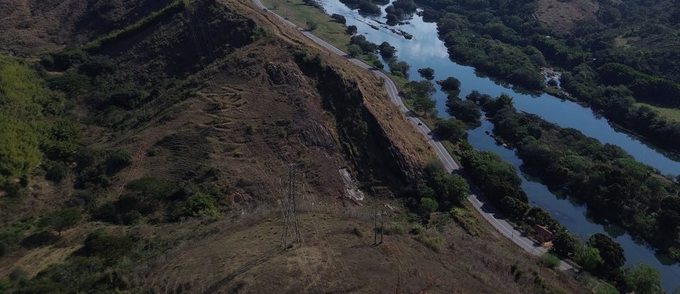 Torre de linha de transmissão existente em encosta de pastagem, com a rodovia e o rio ao fundo