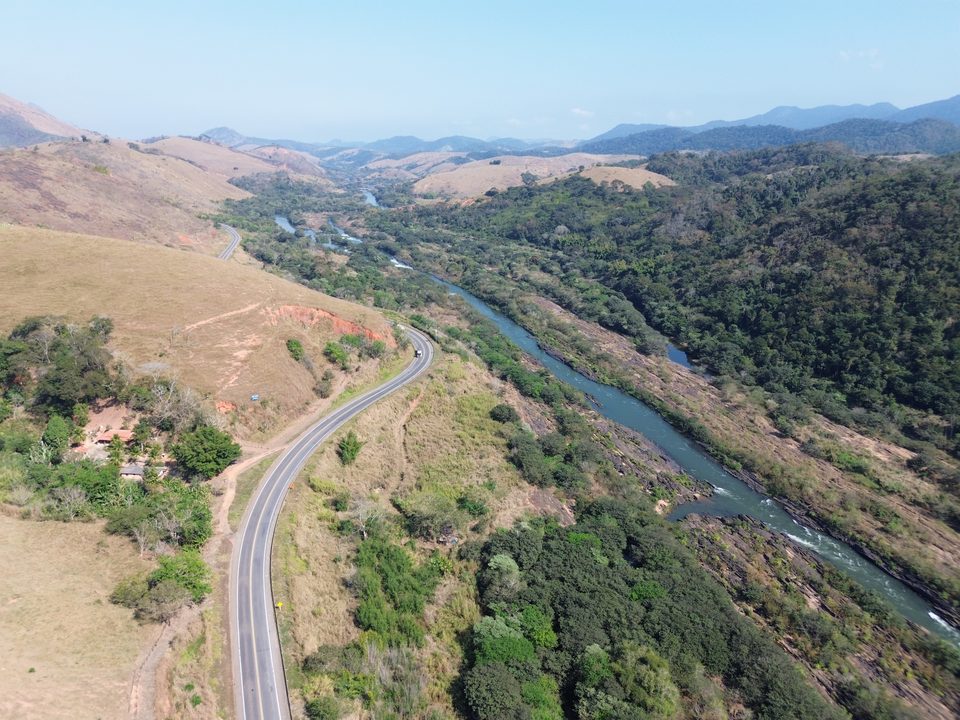 Rodovia sinuosa acompanhando o rio em meio a mata e pastagens
